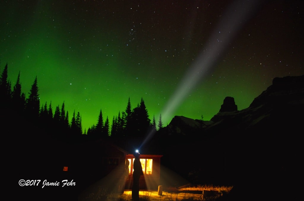 Photographer self-portrait with Northern Lights and cabin in mountains.